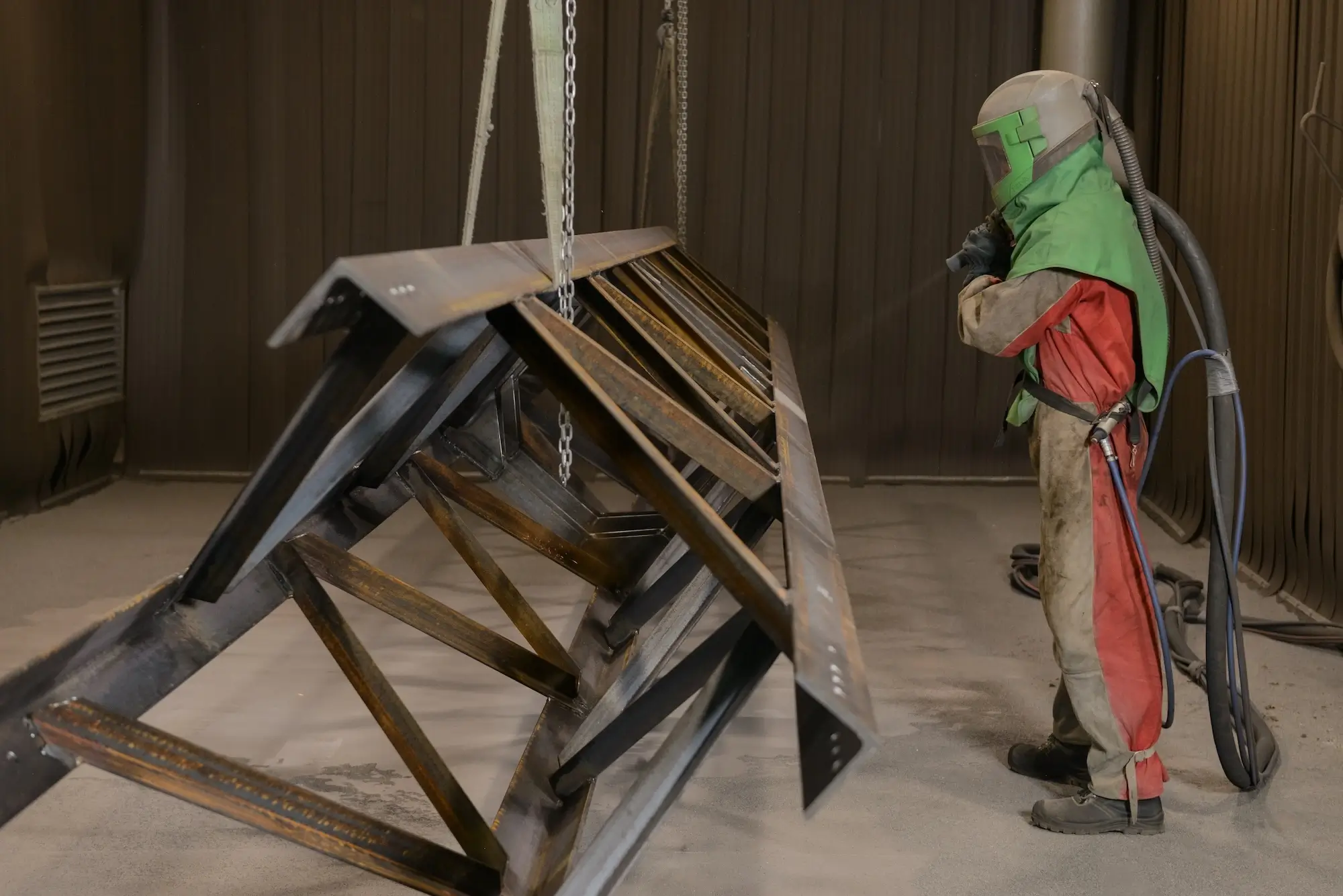 Operator in protective gear performing shot blasting of a large metal frame in a cleaning chamber.