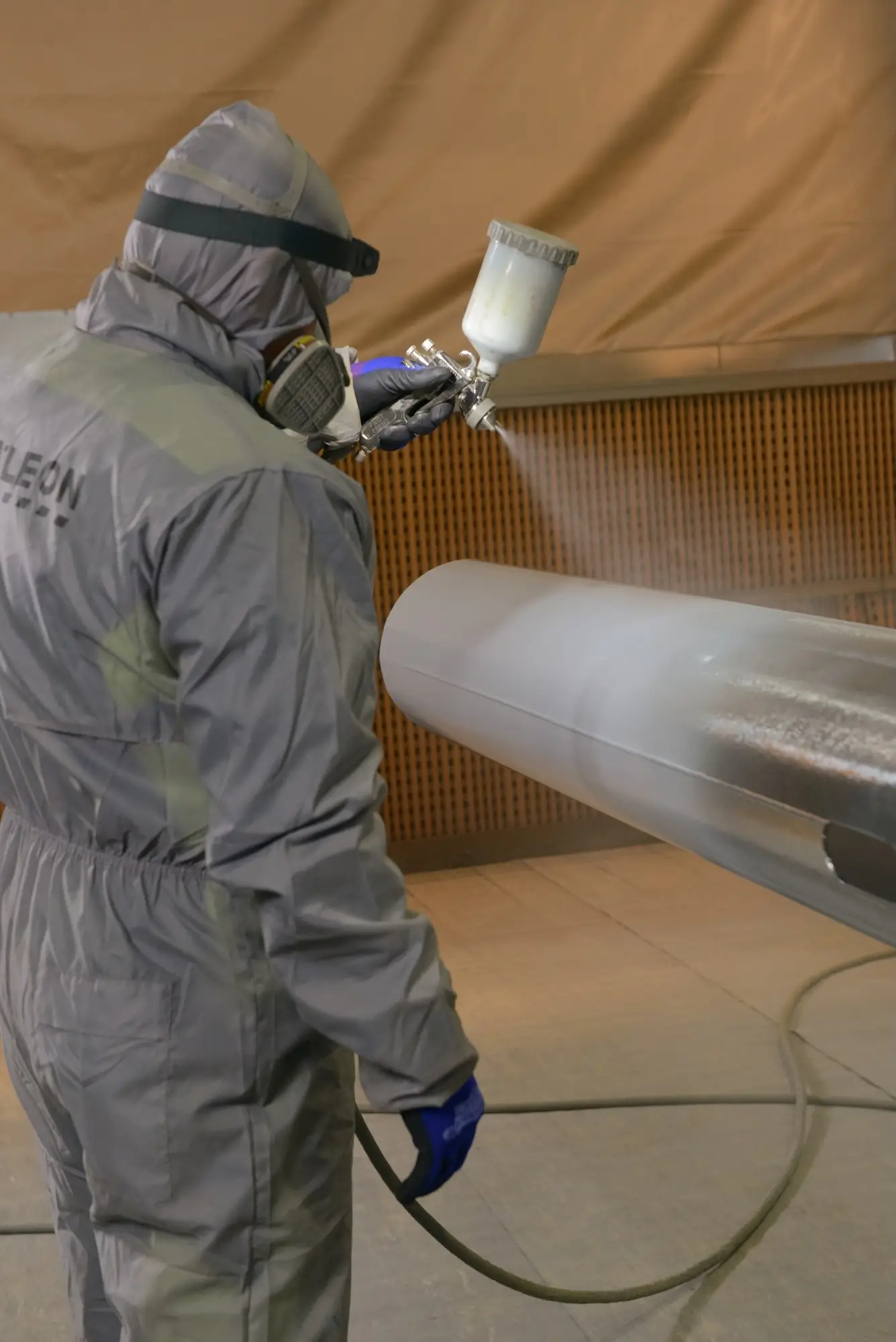 Operator in protective gear performing industrial wet painting of a large metal pipe in a chamber.