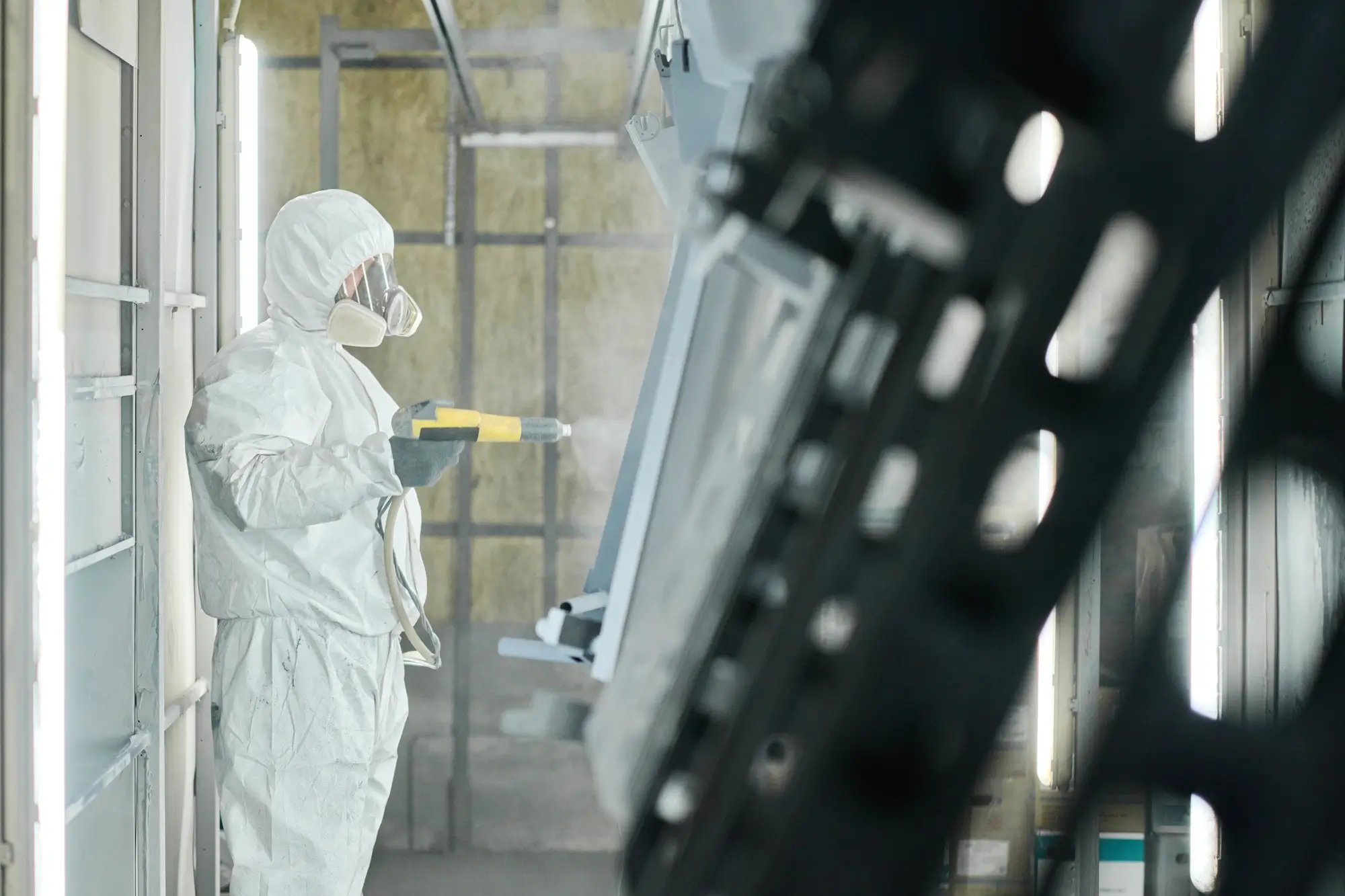 Specialist in protective gear performing manual powder coating of complex metal structures in a chamber.